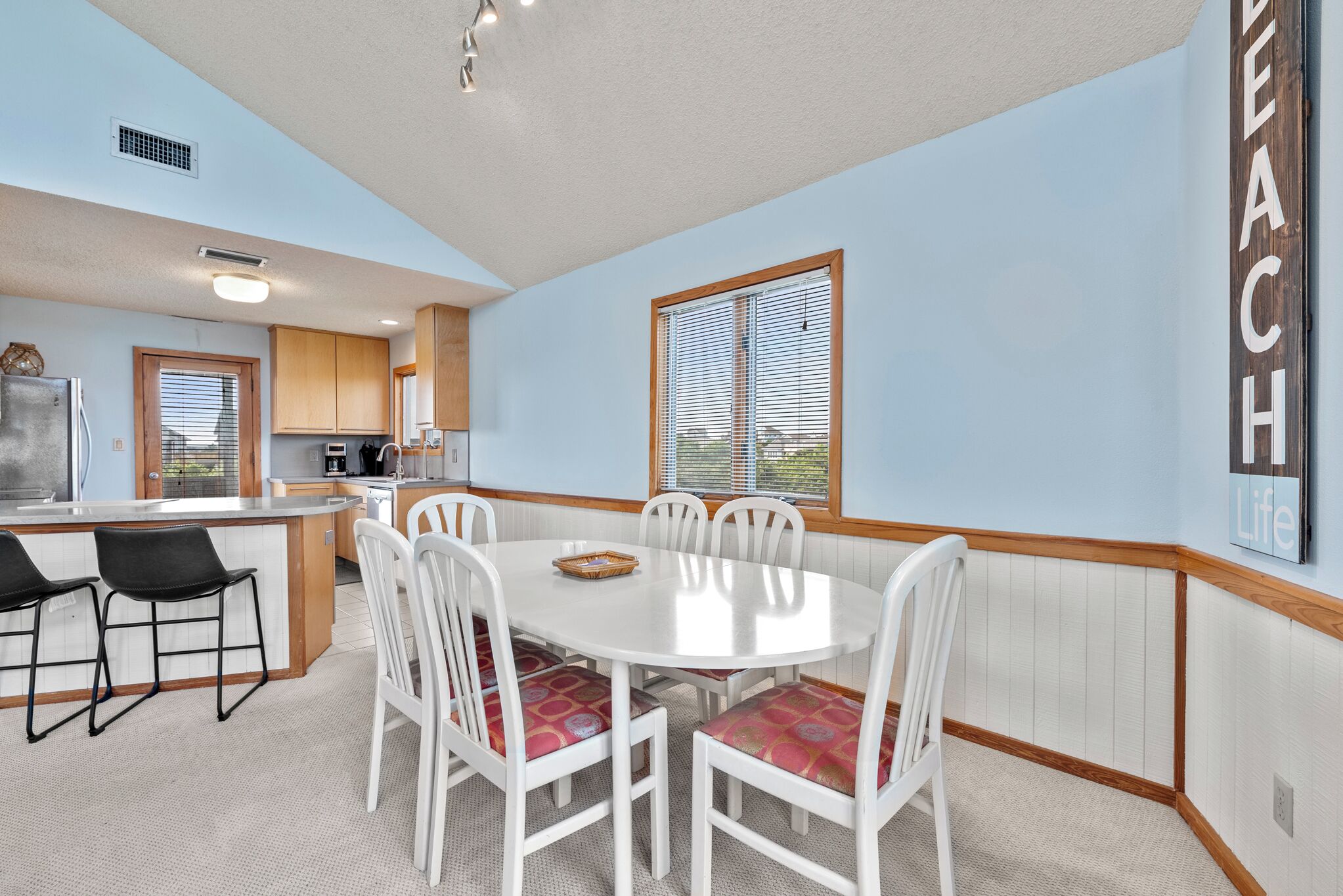 A dining area with a white table and chairs, a kitchen in the background, and a 'Beach Life' sign on the wall.