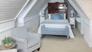 A bedroom with a sloped ceiling, a bed with blue bedding, a white armchair, and a small table with a plant. - 12-Lighthouse Suite at The Castle Inn (Ocracoke)