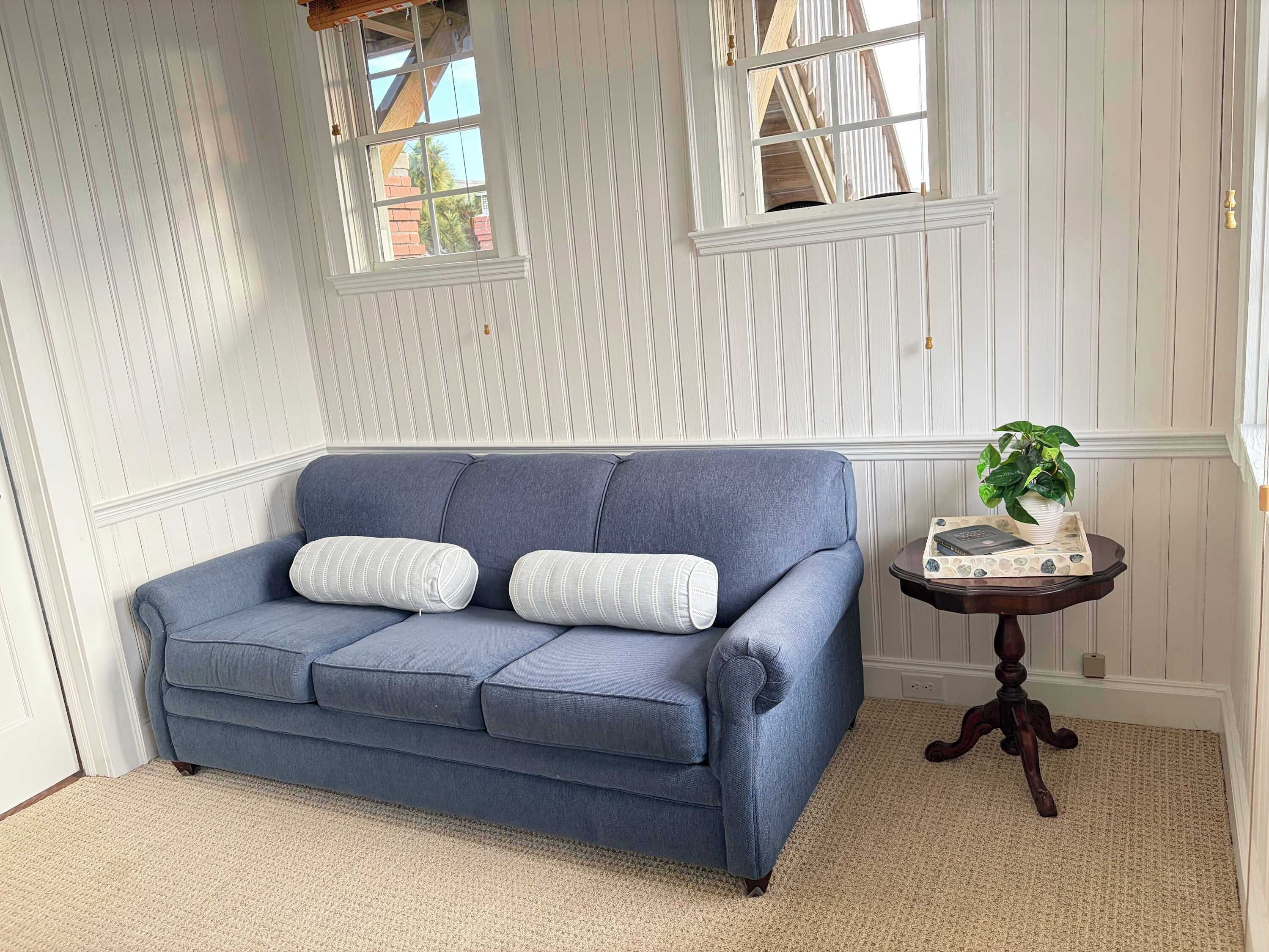 A room with a blue sofa, two white pillows, a small round table with a plant, and a window with a view of a brick building.