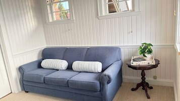 A room with a blue sofa, two white pillows, a small round table with a plant, and a window with a view of a brick building.