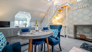 Una sala moderna con una mesa redonda de comedor, sillas tapizadas en azul, un sofá con cojines estampados, una escalera con escalones de madera y una chimenea.
