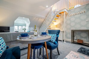 Ein modernes Wohnzimmer mit einem runden Esstisch, blauen Polsterstühlen, einer Couch mit gemusterten Kissen, einer Holztreppe und einem Kamin.