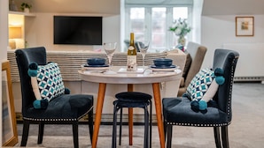Un comedor con una mesa redonda dispuesta para dos personas, que incluye una botella de vino, copas y un plato. Las sillas tienen cojines estampados.