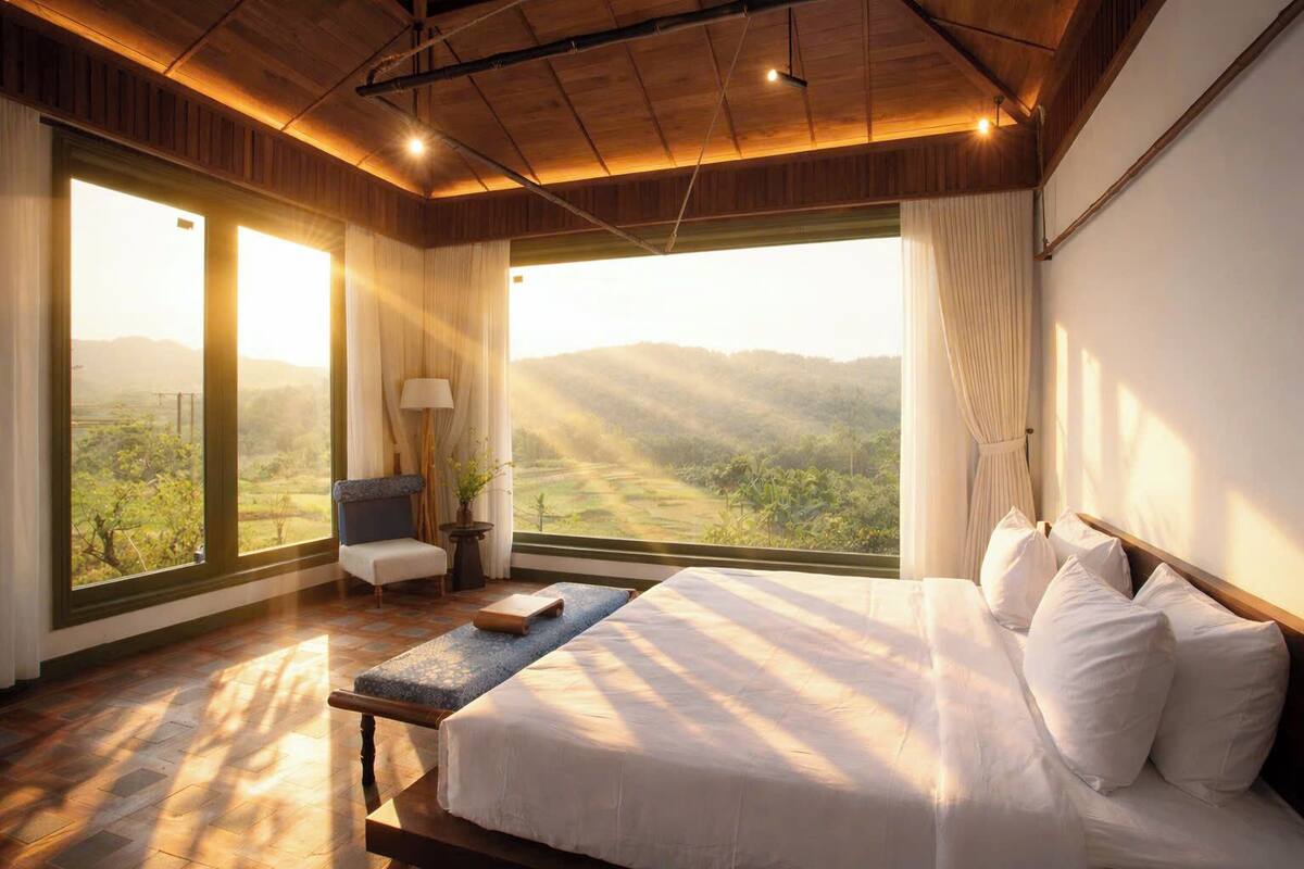 Une chambre avec un grand lit, une chaise, une table et une vue sur un paysage, visible à travers de grandes fenêtres.