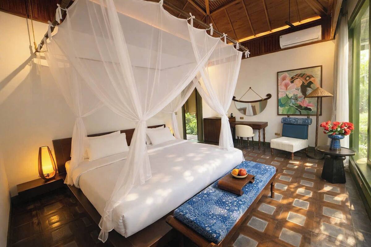 Une chambre à coucher avec un lit à baldaquin, une table de chevet avec une lampe, un banc sur lequel est posé un panier de fruits, et une vue sur l’extérieur.