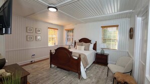 A bedroom with a wooden bed, two bedside tables, a chair, and a television mounted on the wall. - 03-Driftwood Suite at The Castle Inn (Ocracoke)