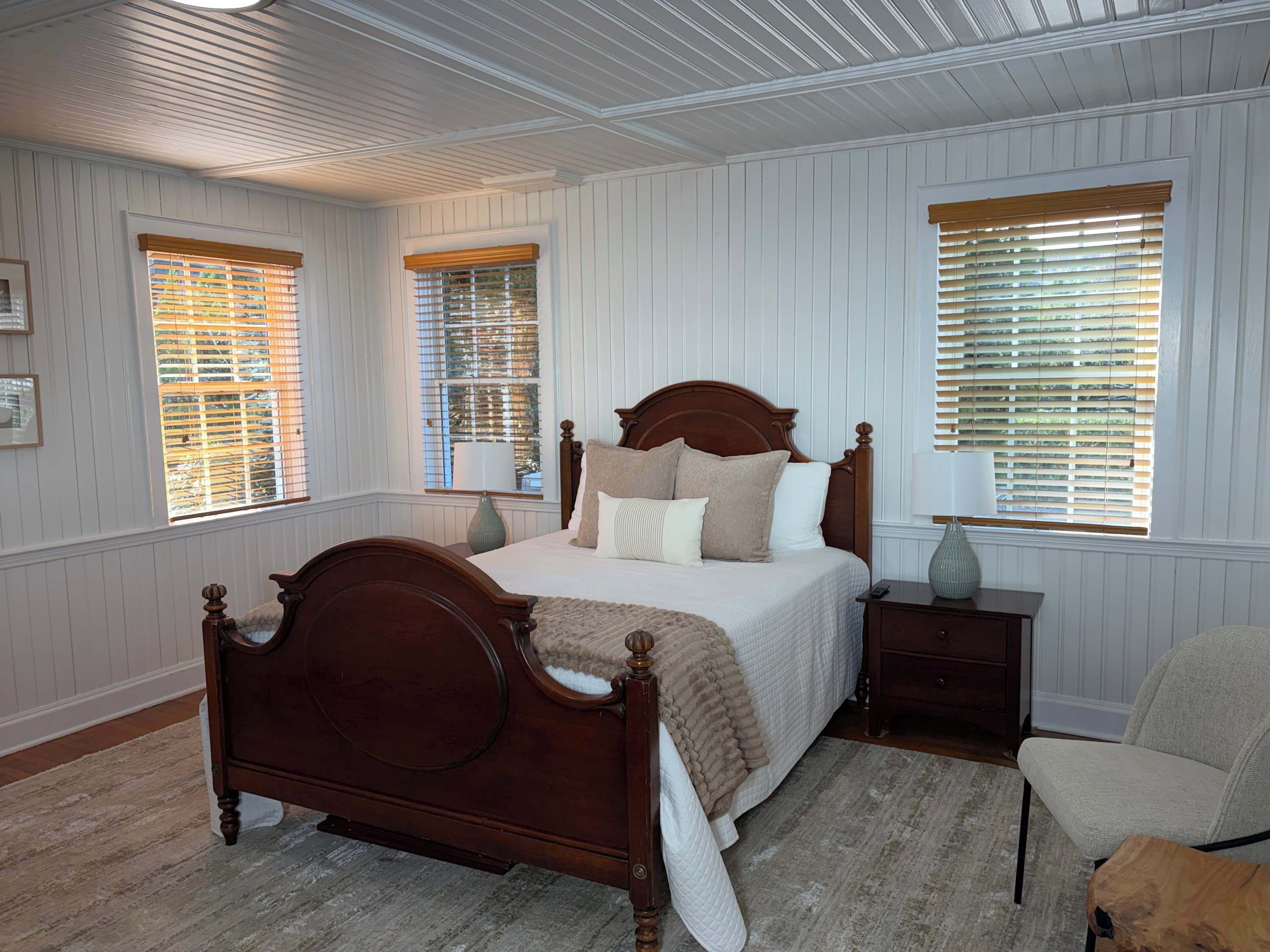 A bedroom with a wooden bed, white walls, and windows with blinds.