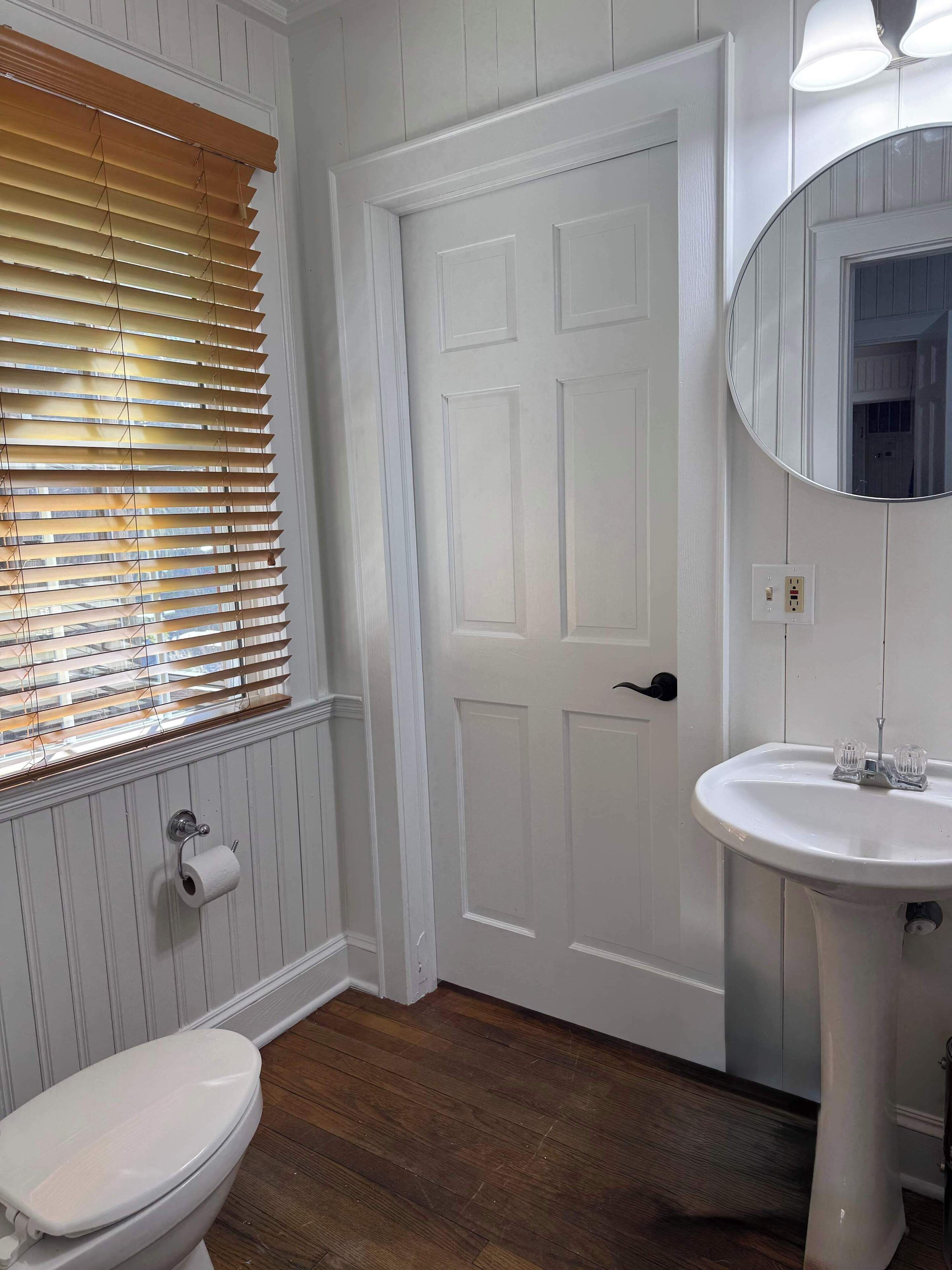 A bathroom with a white door, a toilet, a sink with a mirror, and wooden blinds.