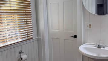 A bathroom with a white door, a toilet, a sink with a mirror, and wooden blinds.