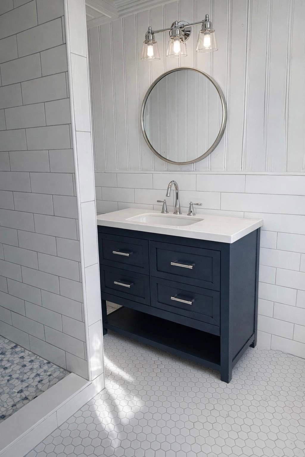 A bathroom with a white tiled wall, a dark blue vanity with three drawers, a round mirror, and a white sink.