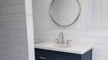 A bathroom with a white tiled wall, a dark blue vanity with three drawers, a round mirror, and a white sink.