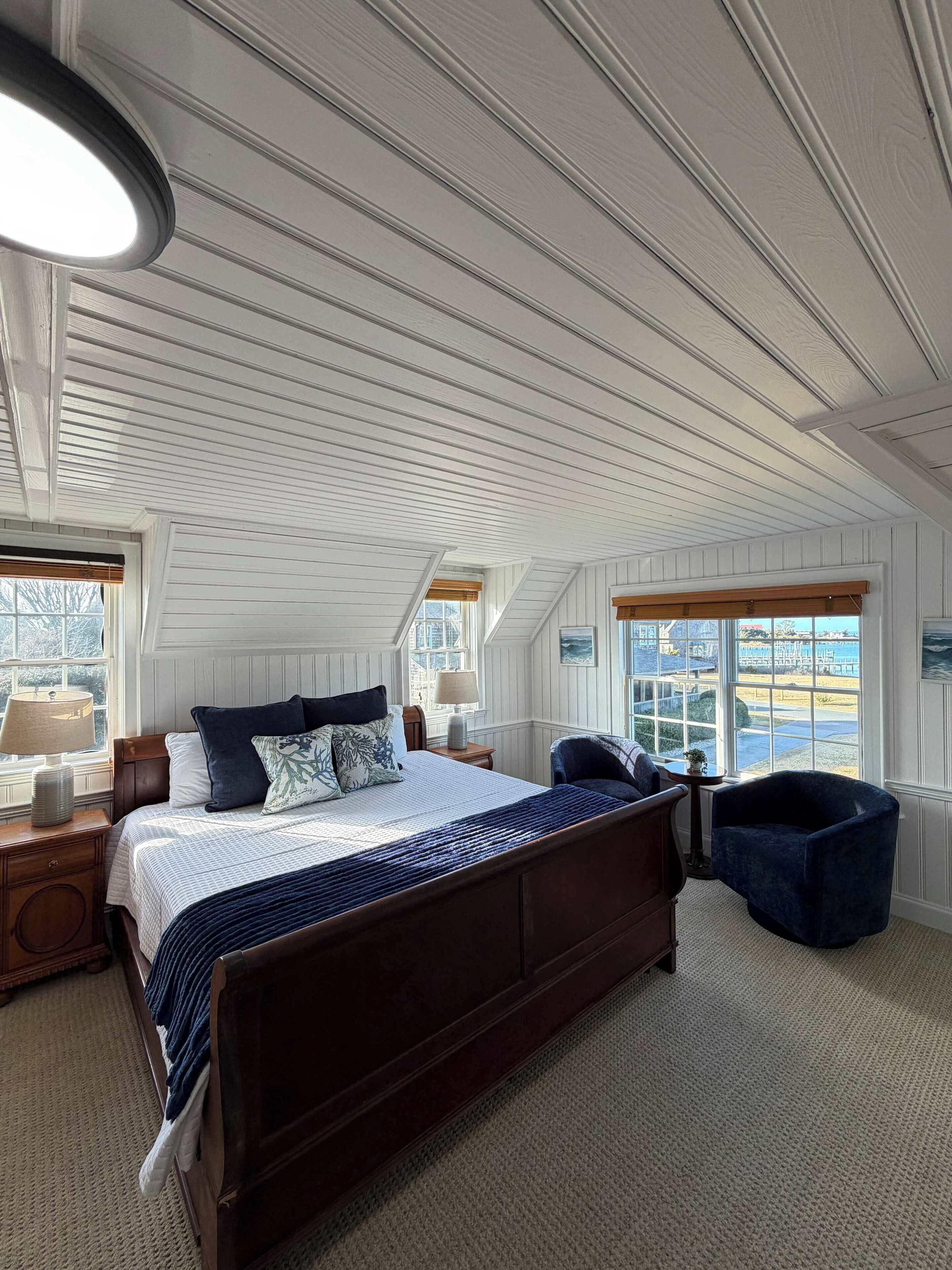 A bedroom with a large bed, a wooden headboard, a nightstand, a lamp, a chair, and a view of the outdoors through a window.