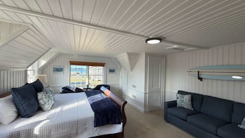 A bedroom with a large bed, a dark-colored sofa, a window with a view of buildings, and a ceiling with wooden beams.