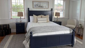 Un dormitorio con una cama azul oscuro, ropa de cama blanca, dos mesitas de noche con lámparas, una silla y un cuadro en la pared.