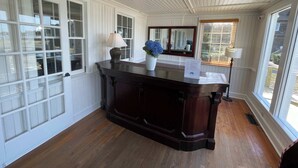 Una habitación con piso de madera, un escritorio de madera oscura, un jarrón blanco con flores azules y una ventana con persianas.