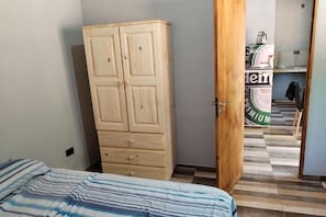 Un dormitorio con cama, un armario de madera y una puerta que da a un pasillo con suelo de baldosas.