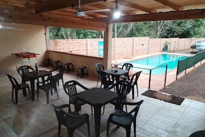 Un patio exterior con mesas y sillas, una piscina y un muro de ladrillo al fondo.