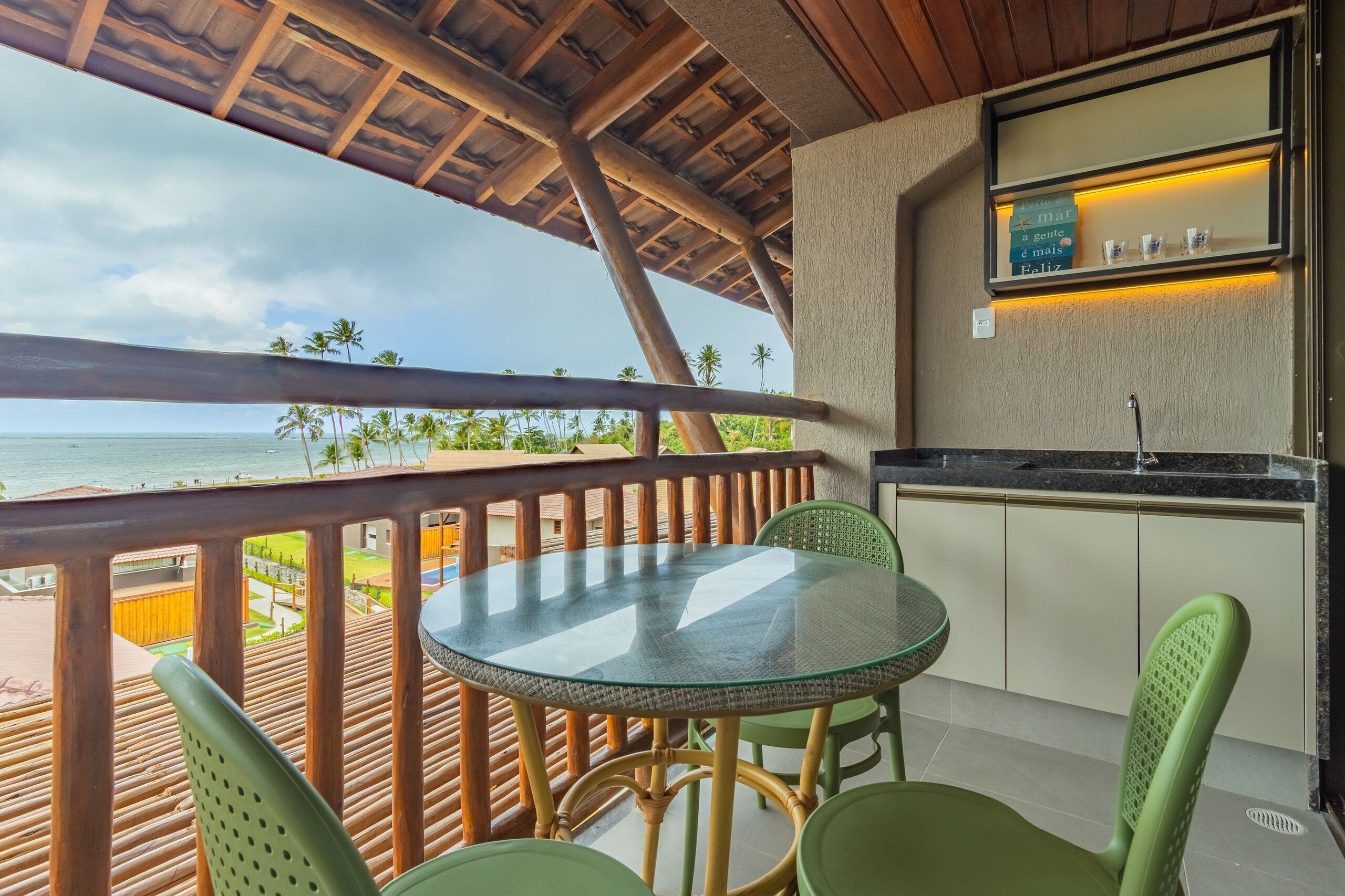 A balcony with a glass table and chairs, overlooking a beach with palm trees and the ocean.