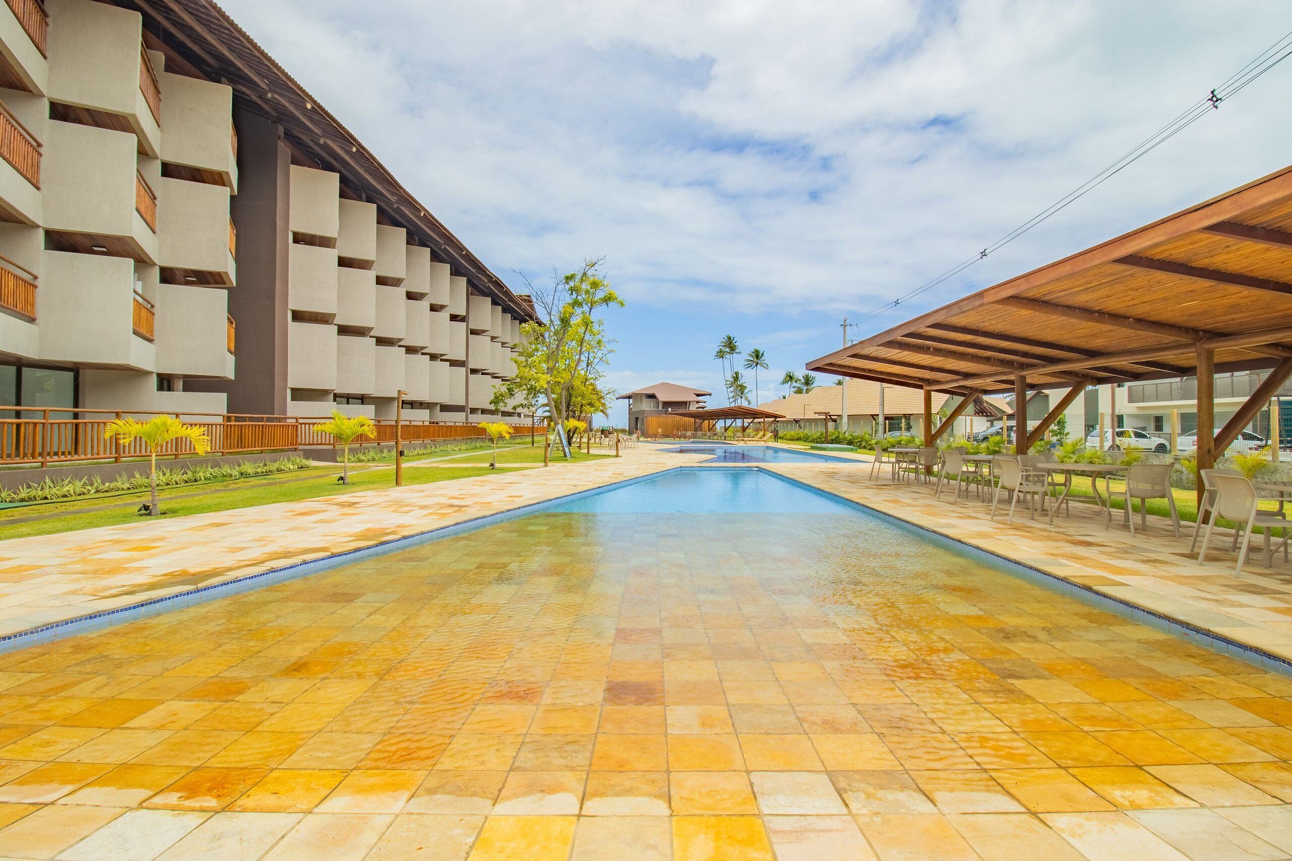 A swimming pool with tiled flooring, surrounded by a covered seating area and a building with multiple balconies.