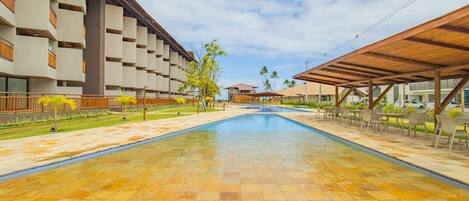 A swimming pool with tiled flooring, surrounded by a covered seating area and a building with multiple balconies.