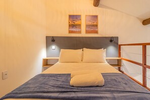 Un lit bien fait, avec une couverture bleue et une serviette pliée, une tête de lit en bois, deux tables de chevet avec des lampes, et deux tableaux encadrés représentant un coucher de soleil sur la mer, accrochés au mur.