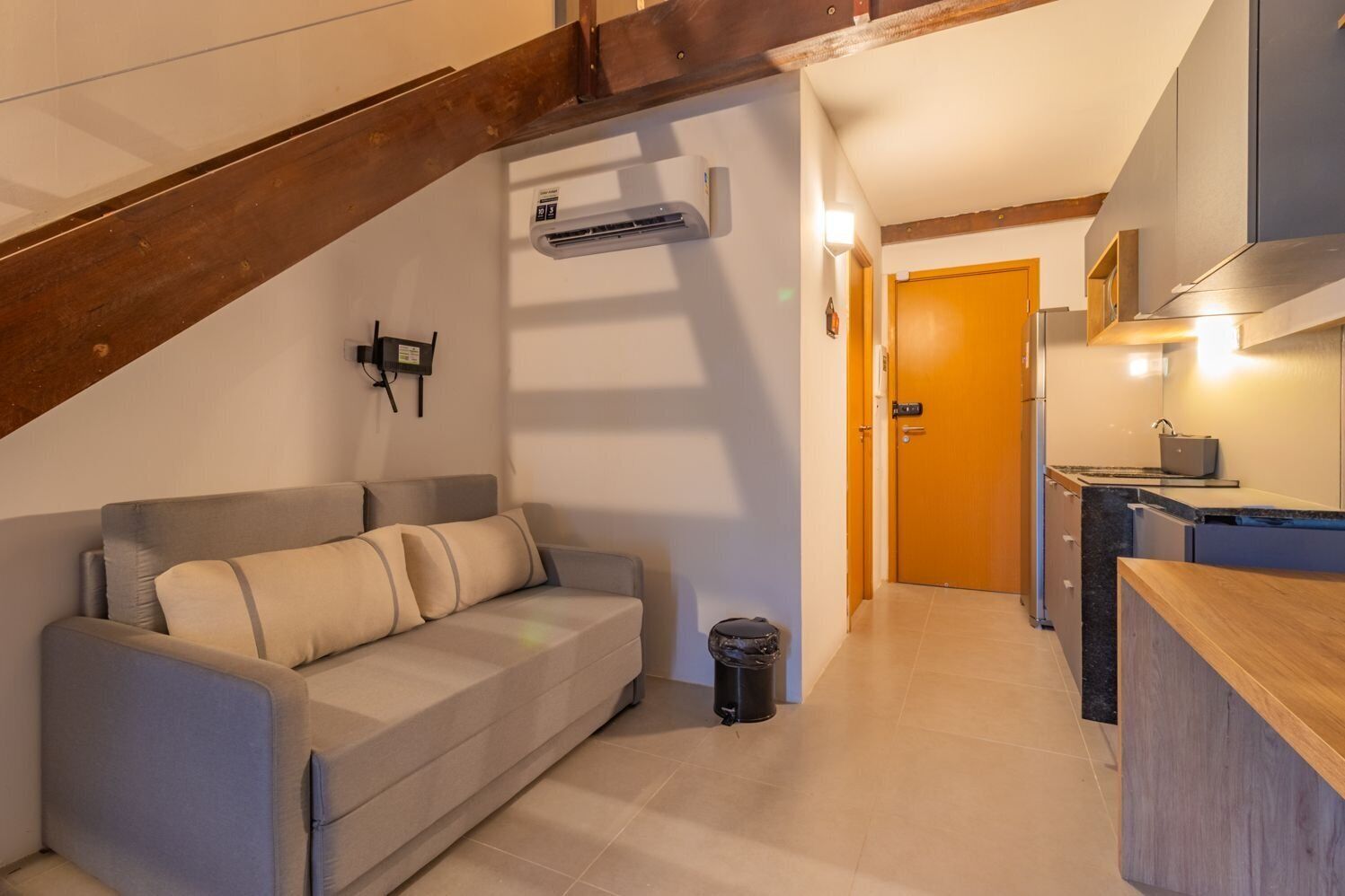 A compact living space with a sofa, a trash bin, a kitchenette, and a wooden staircase.