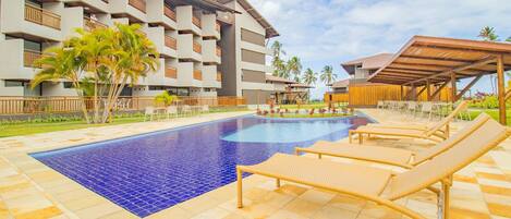 Un hotel con piscina, sillones de descanso y un edificio de varios pisos con balcones.