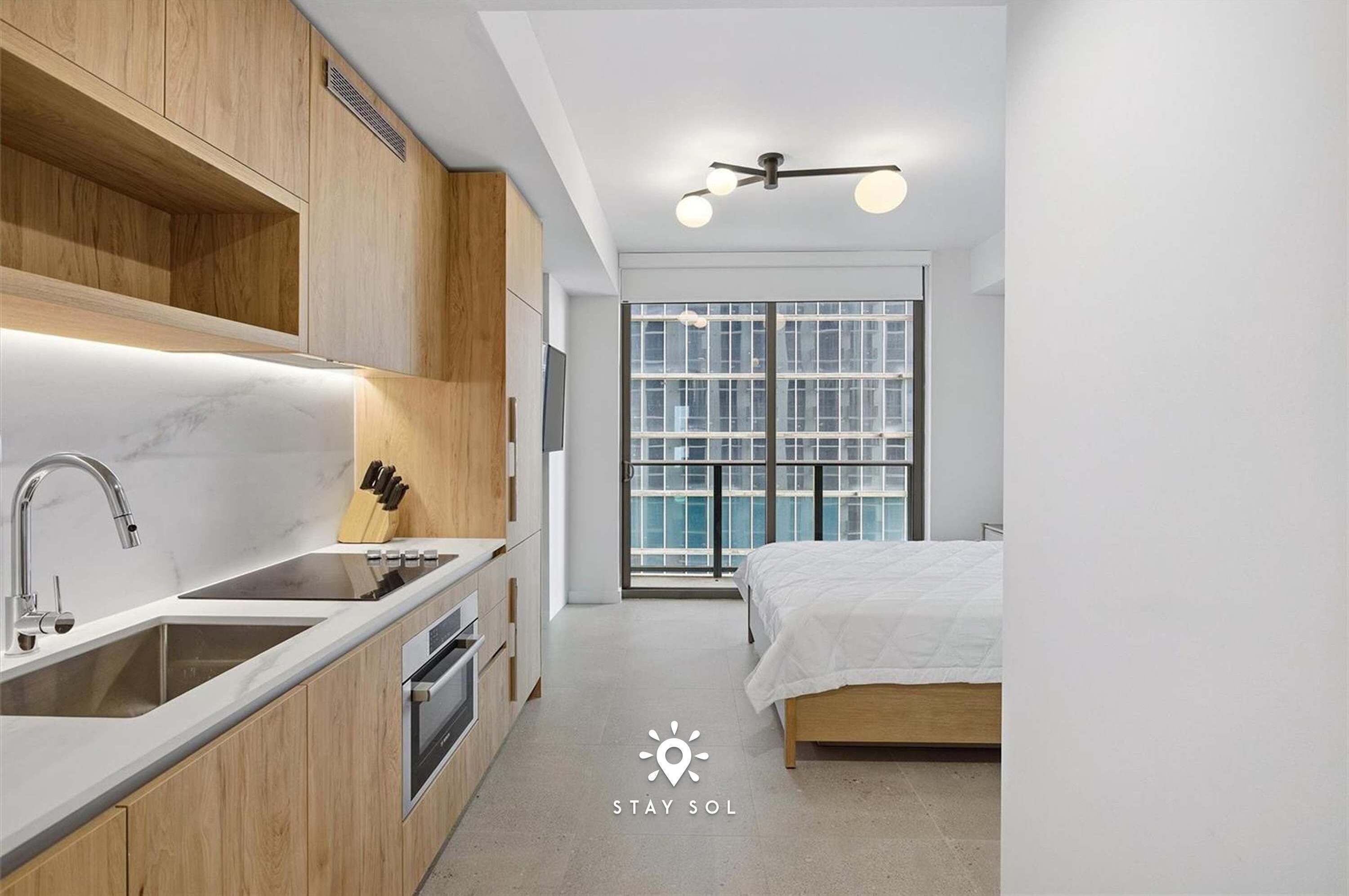 A modern bedroom with a bed, a large window, and a kitchen area with a sink and cabinets.