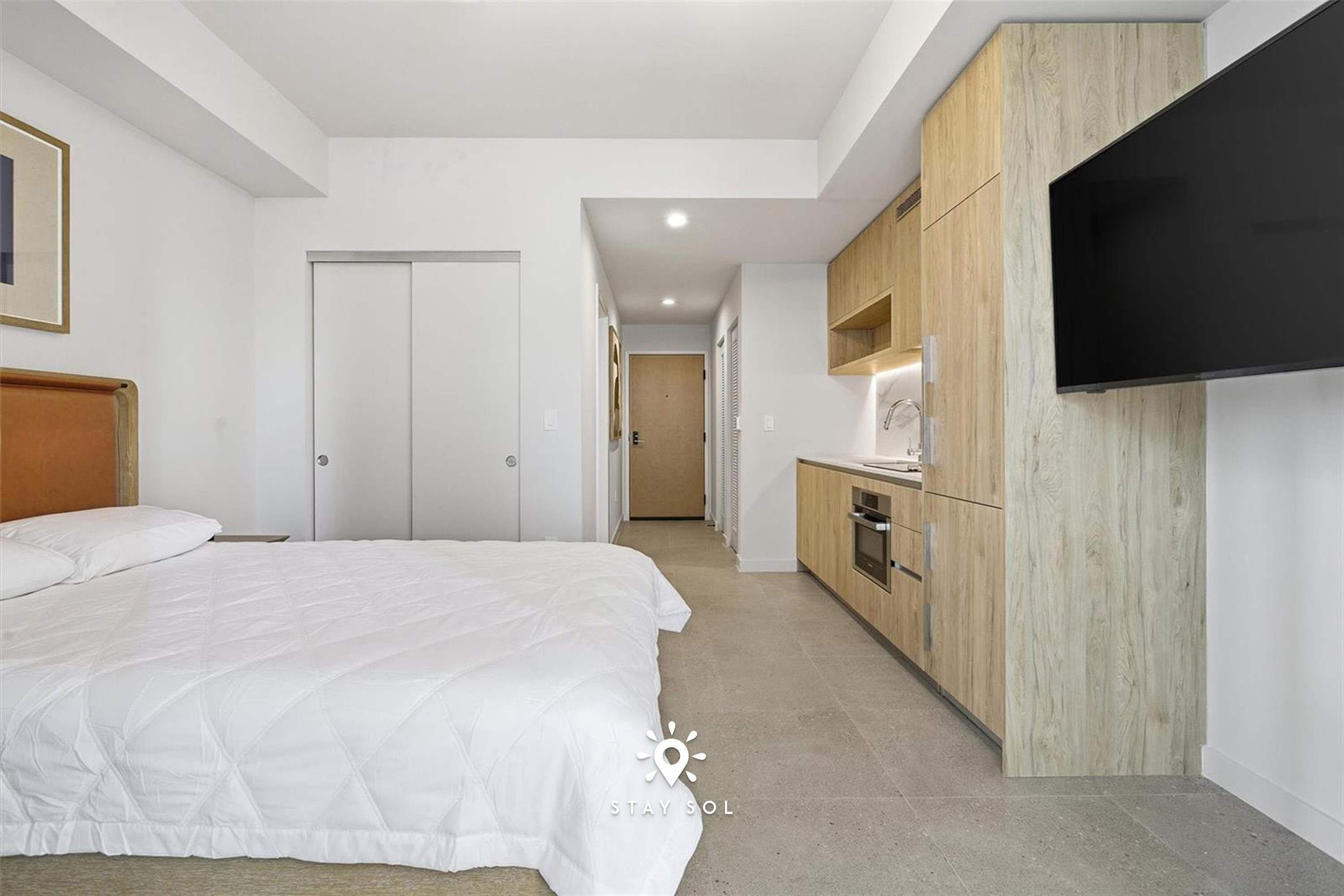 Ein modernes Hotelzimmer mit einem großen Bett, einer Küchenzeile, einem an der Wand montierten Fernseher und einer Tür zu einem weiteren Raum.