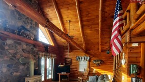 A cozy cabin interior with a stone wall, wooden beams, a fireplace, a comfortable sofa, and an American flag. - A peaceful getaway awaits at this beautiful Log Home located on Sodus Bay (Wolcott)