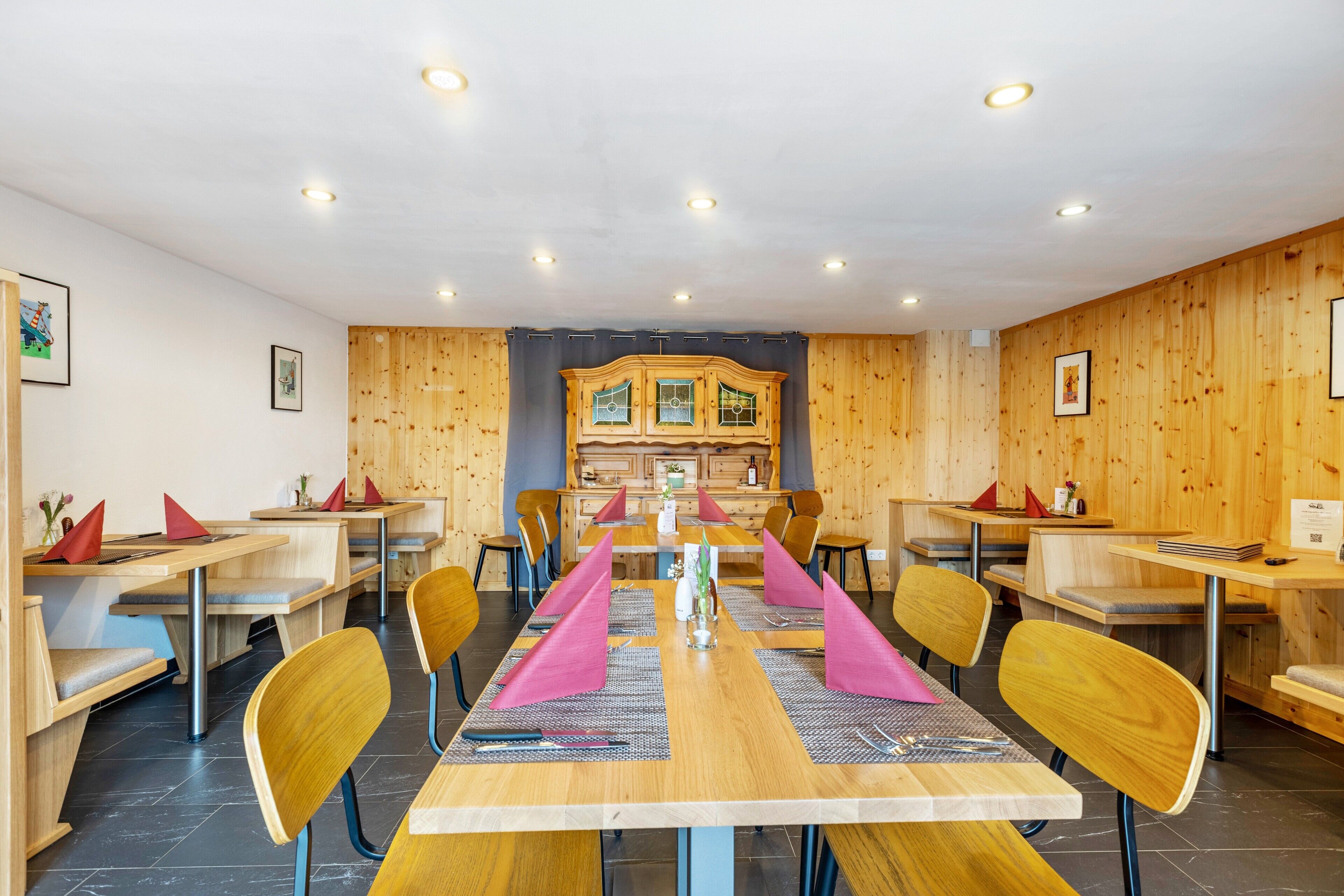 A dining area with wooden panel walls, a central table with pink napkins, and a kitchen in the background.