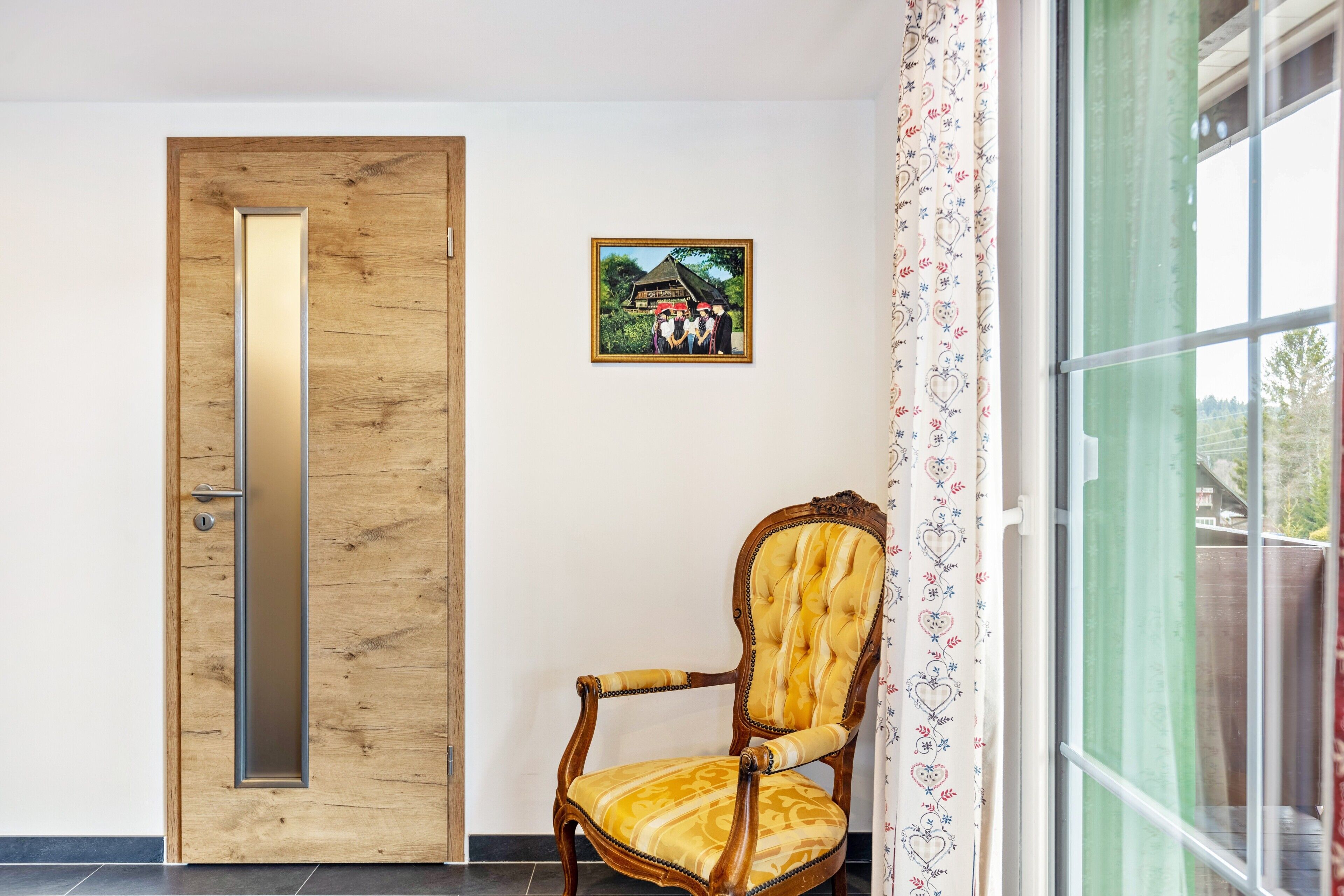 A wooden door with a vertical mirror, a framed picture on the wall, a chair with a yellow cushion, and a window with patterned curtains.