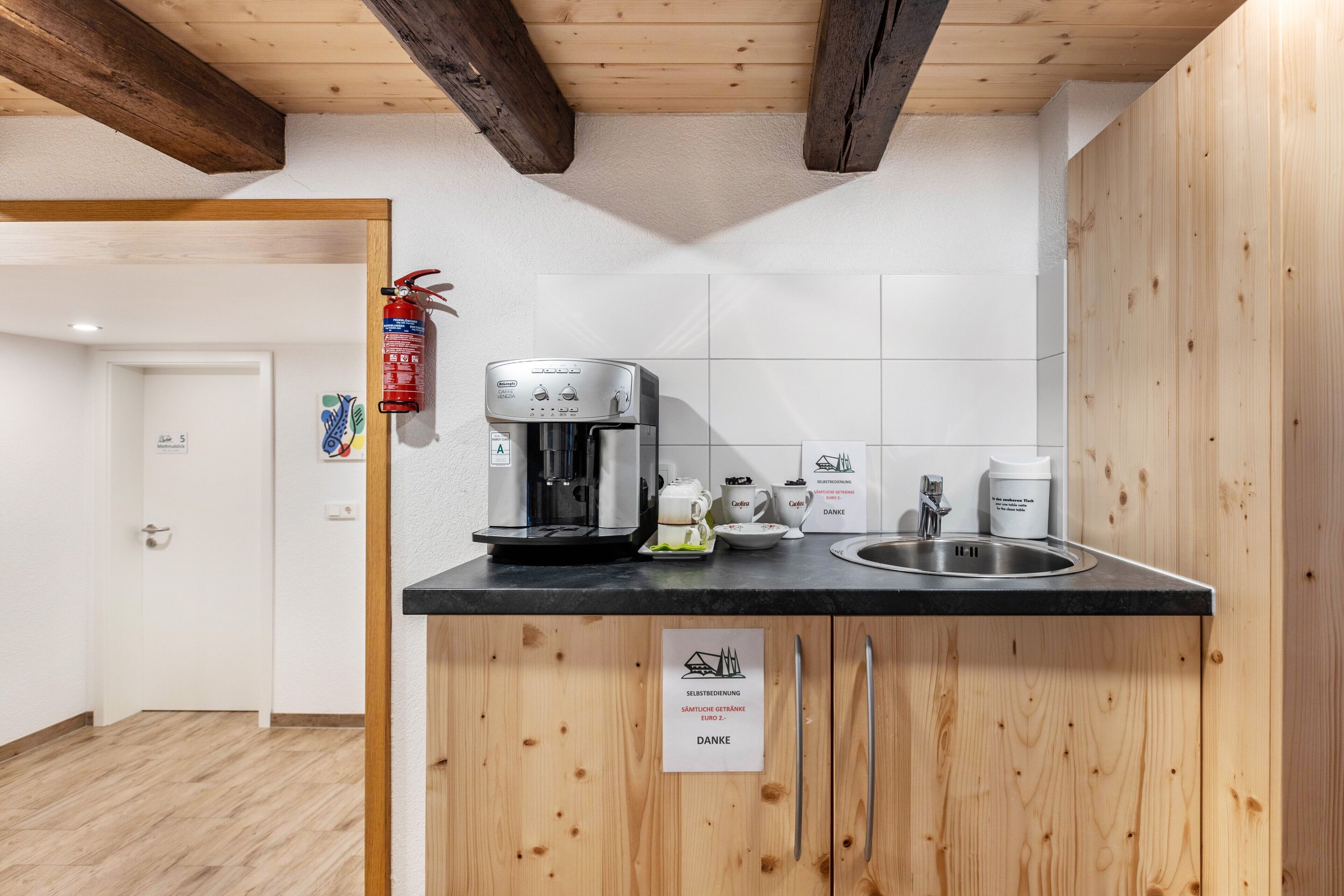 Ett litet kök med en kaffebryggare, en diskho och en skylt som visar att frukosten är avsedd för självbetjäning.
