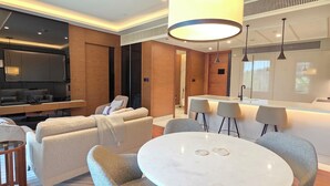 Ein modernes Hotelzimmer mit einem runden Esstisch, grau gepolsterten Sesseln, einer großen Pendelleuchte, einem Flachbildfernseher und einer Holzwand als Gestaltungselement.