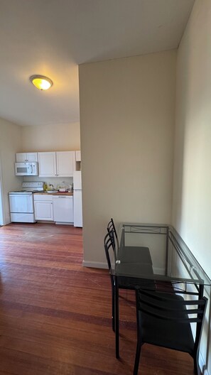 A room with a dining table and chairs, a kitchen area with white cabinets and appliances, and a wooden floor. - Updated 1BR apt, 1st floor, near T, free parking (Malden)