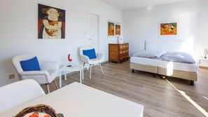 Ein moderner, gut beleuchteter Raum mit einem Bett, zwei weißen Stühlen, einem kleinen Tisch und Bildern an den Wänden.