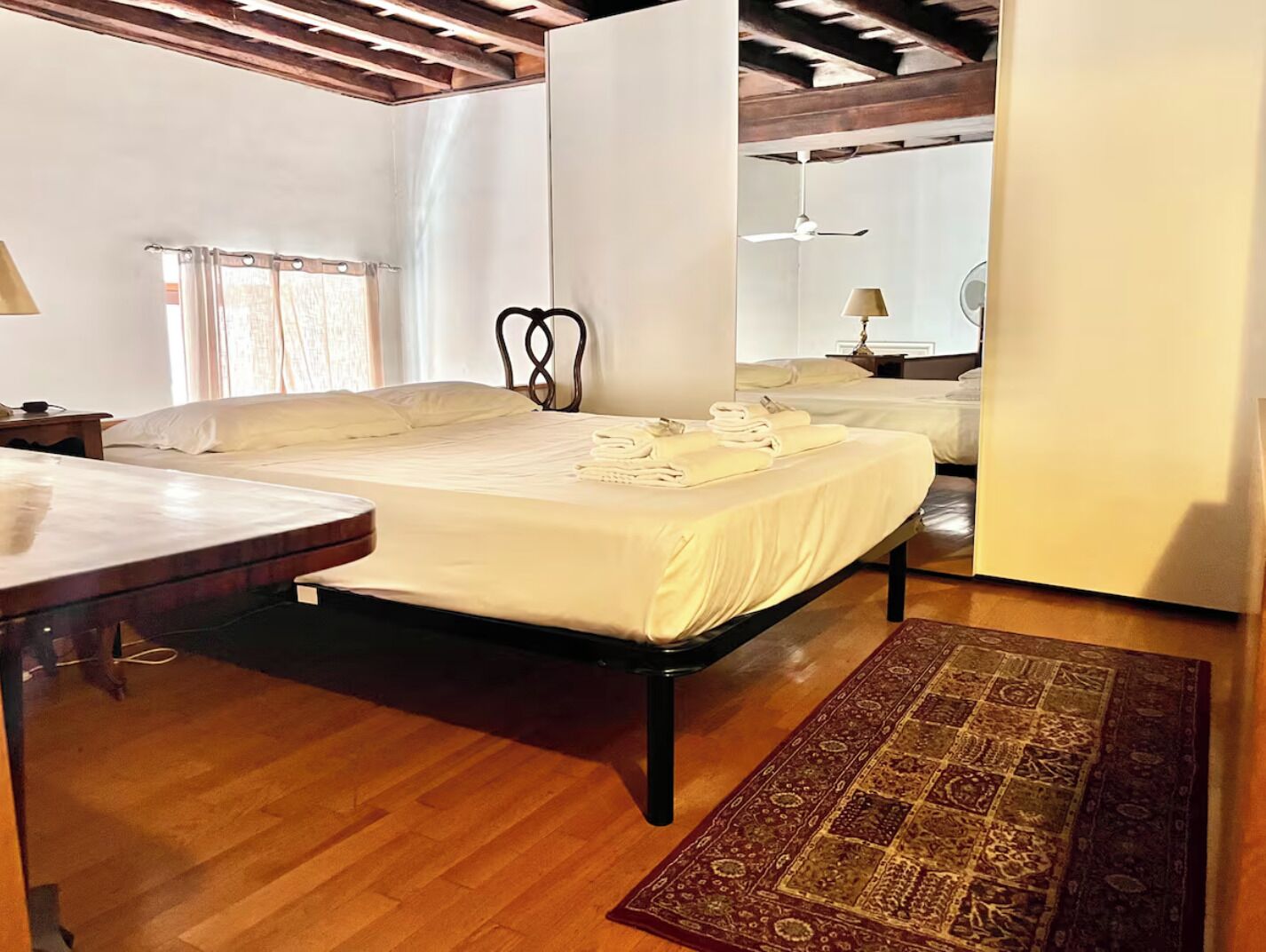A hotel room with a wooden floor, a bed with white linens, a bedside table, a lamp, and a patterned rug.