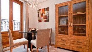 Een eetruimte met een tafel en stoelen, een kroonluchter, een grote houten kast met glazen deuren en een raam met uitzicht op gebouwen.