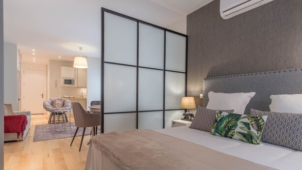 Un dormitorio moderno con una cama grande, una zona de estar independiente con una silla y una mesa, y una kitchenette al fondo.