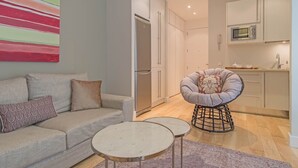 Una sala de estar moderna con un sofá, una mesa de centro y una silla original. Se ve una cocina al fondo con un microondas y armarios.