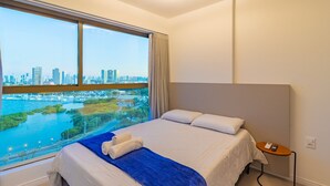 A hotel room with a large window offering a city view, a neatly made bed, and a small bedside table with a phone. - Parque do Cais #1210 - Apt With a View by Carpediem (Recife)