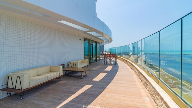 A modern outdoor lounge area with a curved glass railing, a sofa, and a table. - Parque do Cais #1210 - Apt With a View by Carpediem (Recife)