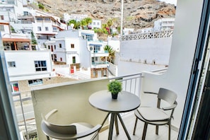 A balcony with two chairs and a small table, overlooking a coastal town with white buildings and a rocky hillside. - Casa Arno - Lovely Modern Apartment In Puerto De Mogan (Lomo Quiebre)