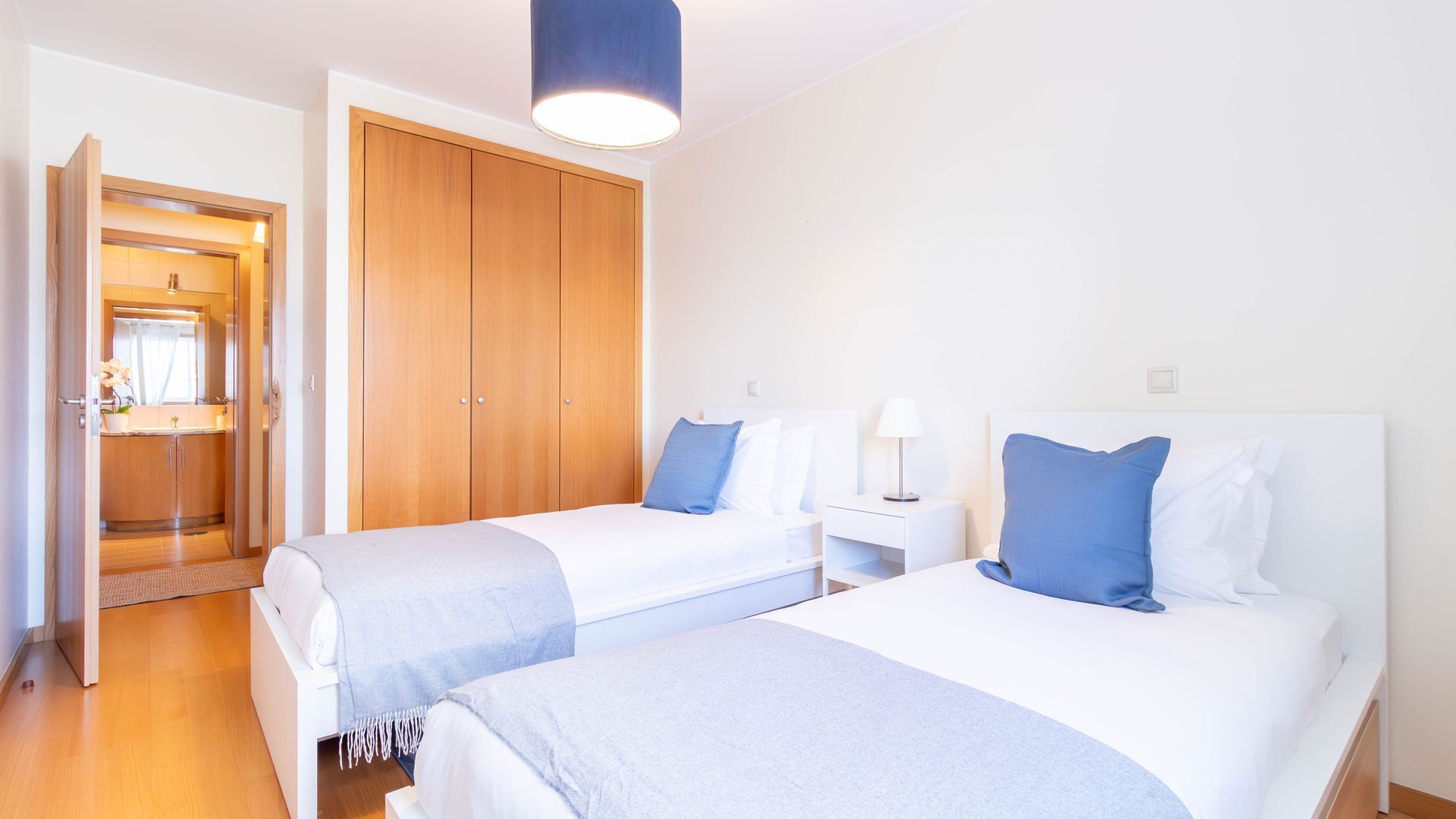 Une chambre d’hôtel avec deux lits, une armoire en bois, une salle de bain avec un miroir et un luminaire bleu au plafond.