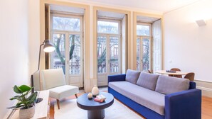 A modern living room with a blue sofa, a white armchair, a round coffee table, and a floor lamp. Large windows provide a view of the outside. - B. Arts 1F by LovelyStay (Porto)
