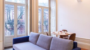 A room with a blue sofa, a round table with a few items, and large windows with a view of buildings and a balcony. - B. Arts 1F by LovelyStay (Porto)