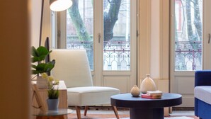 A room with a white chair, a round table with a vase and books, and a view through a window with a metal gate. - B. Arts 1F by LovelyStay (Porto)