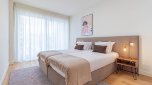 Ein Doppelbett mit Kopfteil, ein Nachttisch mit Lampe und ein großes Fenster mit durchsichtigen Vorhängen.