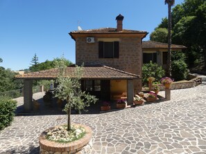 Un bâtiment en pierre avec un toit en tuiles, un petit jardin agrémenté de plantes en pot et une cour dallée de pierres.