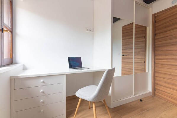 A modern bedroom with a white desk, a laptop, a grey chair, and a wooden wardrobe.